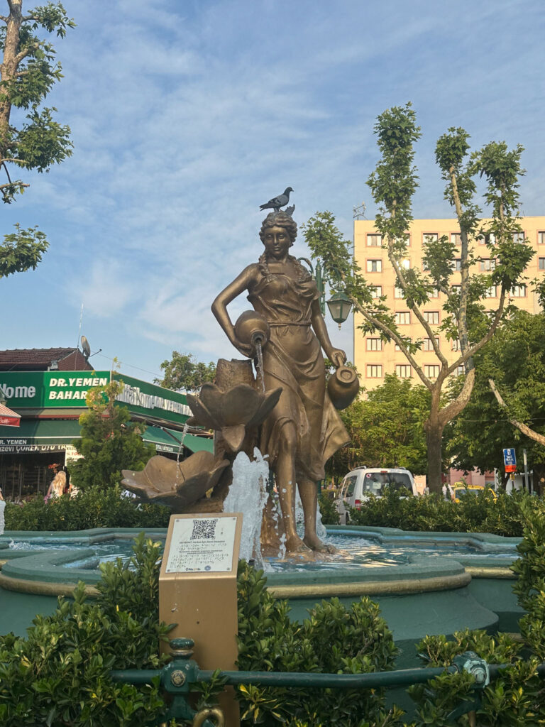 Bronze fountain statue of a woman pouring water in a city square with trees and buildings around. 