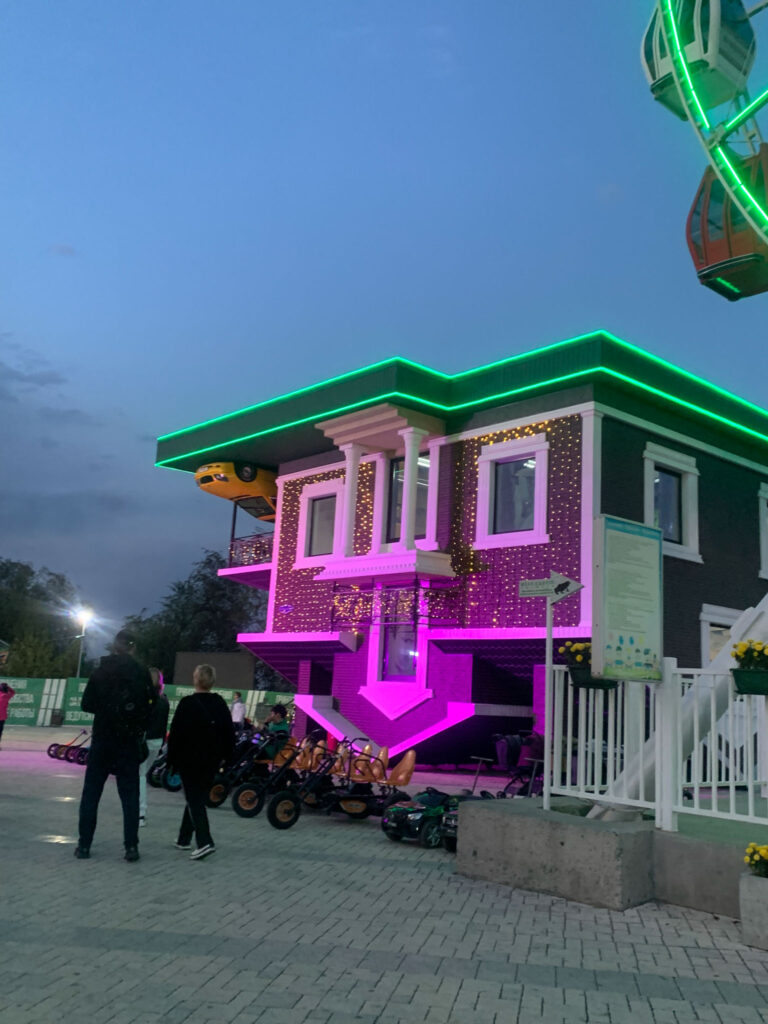 Neon-lit upside-down house attraction with visitors walking nearby at an amusement park during evening.